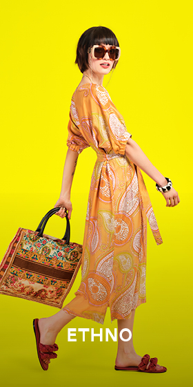 Eine stylische Frau in einem gemusterten gelben Kleid und Sonnenbrille posiert vor einem leuchtend gelben Hintergrund. Sie trägt eine bunt gemusterte Tasche und rote Sandalen. Unten steht der Text „ETHNO“.