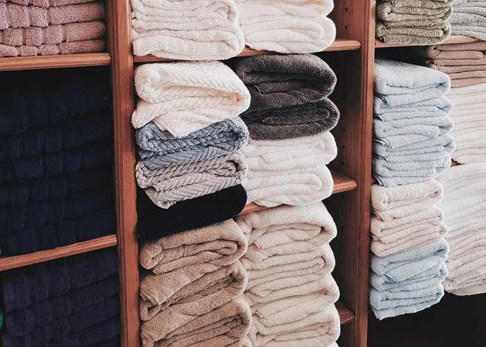 Stapel ordentlich gefalteter Handtücher in verschiedenen Farben, darunter Weiß, Beige, Grau und Blau, sind auf Holzregalen angeordnet.