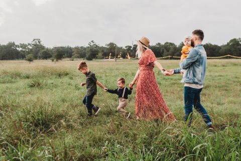 Eine fünfköpfige Familie geht über eine Wiese. Die Mutter, die ein rotes Kleid und einen Hut trägt, hält die beiden kleinen Kinder an den Händen. Der Vater, in einer Jeansjacke, trägt ein Kleinkind. Im Hintergrund sind Bäume unter einem bewölkten Himmel zu sehen.