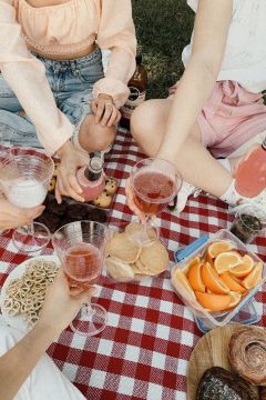 Vier Personen sitzen auf einer rot-weiß karierten Picknickdecke. Sie halten Getränke in verschiedenen Gläsern. Auf der Decke liegen Snacks wie Kekse, Orangenscheiben, Brezeln und Brownies. Die Atmosphäre ist entspannt und locker.