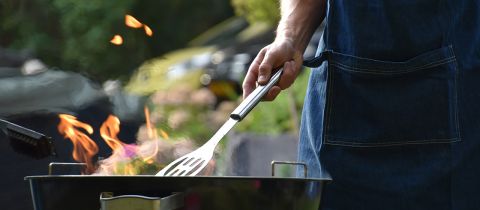 Eine Person mit Schürze grillt im Freien und hält einen Spatel über offene Flammen. Im Hintergrund sind verschwommenes Grün und ein geparktes Fahrzeug zu sehen.