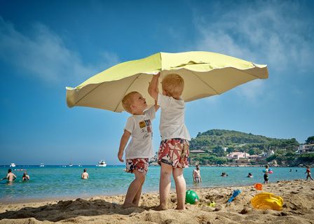 Zwei Kleinkinder in passenden Outfits stehen unter einem großen Sonnenschirm an einem sonnigen Strand. Im Hintergrund sind das Meer und mehrere schwimmende Menschen zu sehen, zusammen mit grünen Hügeln und einem klaren blauen Himmel.