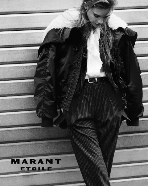 Schwarz-Weiß-Foto einer Frau, die eine Jacke mit Schurwollkragen, eine Rüschenbluse und eine Nadelstreifenhose trägt und sich an eine Metallwand lehnt. Der Text "MARANT ETOILE" steht unten links.