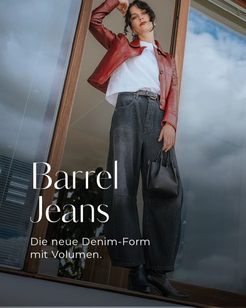 Eine Frau in weit geschnittenen Röhrenjeans, weißem T-Shirt, roter Lederjacke und Stiefeln steht vor einem Glasfenster und hält eine schwarze Handtasche. Der Text lautet: "Barrel Jeans. Die neue Denim-Form mit Volumen.
