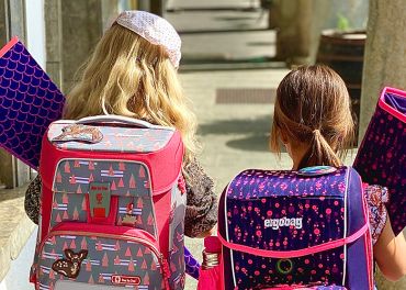 Zwei Kinder gehen auf einem sonnigen Weg und tragen jeweils bunte Rucksäcke und Ordner. Das linke Kind hat einen rosa Rucksack mit Baummustern und das rechte Kind einen dunklen Rucksack mit violetten Mustern. Sie gehen in Richtung eines schattigen Bereichs.