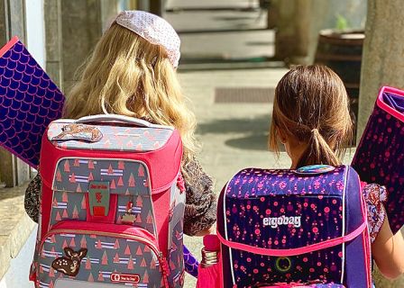 Zwei Kinder gehen auf einem sonnigen Weg und tragen jeweils bunte Rucksäcke und Ordner. Das linke Kind hat einen rosa Rucksack mit Baummustern und das rechte Kind einen dunklen Rucksack mit violetten Mustern. Sie gehen in Richtung eines schattigen Bereichs.