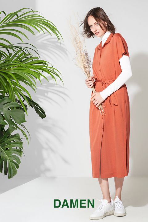Eine Frau steht da und hält getrocknete Blumen in der Hand. Sie trägt ein orangefarbenes Kleid über einem weißen Langarmshirt. Sie lächelt leicht. Auf der linken Seite des Bildes sind große grüne Blätter zu sehen. Unten steht das Wort „DAMEN“.