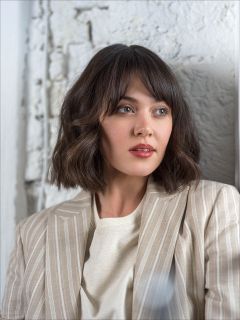 Frau mit kurzen, gewellten braunen Haaren, die einen beigen Nadelstreifenblazer und ein weißes Oberteil trägt und zur Seite gegen eine strukturierte weiße Wand blickt.