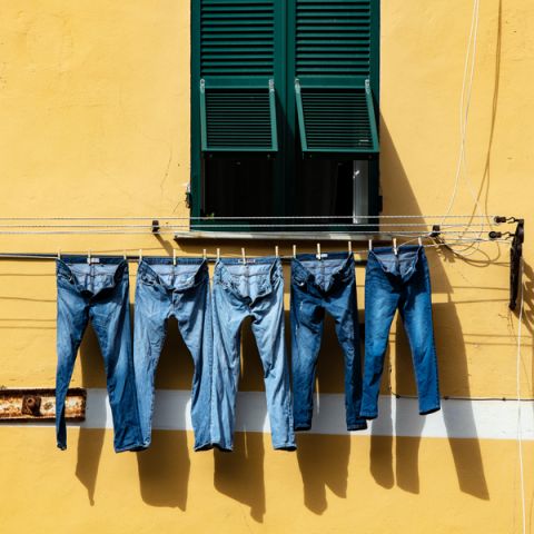 Fünf Paar Bluejeans hängen auf einer Wäscheleine vor einer gelben Wand. Darüber ist ein Fenster mit grünen Fensterläden teilweise geöffnet und Sonnenlicht wirft Schatten auf die Wand.