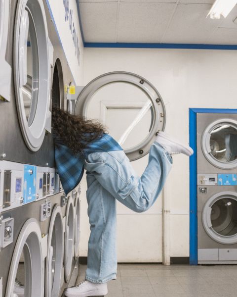 Eine Person in einem blau karierten Hemd und Jeans lehnt sich in einem Waschsalon an eine Waschmaschine. Ihr rechtes Bein ist zum Balancieren nach hinten angehoben. Der Raum verfügt über mehrere Waschmaschinen und Trockner in blau-weißen Farbtönen.
