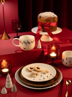 Eine festlich gedeckte Tafel mit einem großen Kuchen, der mit einer roten Schleife umwickelt und mit Puderzucker bestäubt ist, auf einem Ständer. Es gibt Kekse auf Tellern, Themenbecher, Kerzen, rot-weißen Christbaumschmuck und einen tiefroten Hintergrund.