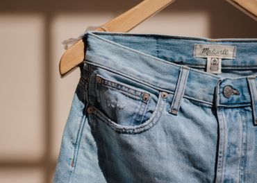 Nahaufnahme einer hellblauen Jeans, die auf einem hölzernen Kleiderbügel hängt. Die Jeans hat am Hosenbund ein Lederetikett mit dem Markennamen „Madewell“ und ist in warmes Sonnenlicht getaucht, wodurch ihre verblasste Textur und Nähte hervorgehoben werden.