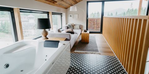 Ein gemütliches Loftzimmer verfügt über ein weißes Sofa, einen kleinen Holztisch und eine schwarze Lampe. Neben einem gemusterten Boden steht eine Badewanne. Große Fenster und eine Glastür öffnen sich zu einem Balkon und bieten einen malerischen Blick auf Bäume und Hügel.
