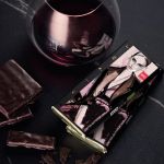 Ein Glas Rotwein steht neben einem dunklen Schokoladenriegel mit einer stilvoll illustrierten Frau auf der Verpackung. Mehrere Schokoladenstücke sind auf einer dunklen Fläche in der Nähe verstreut.