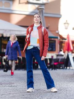 Eine lächelnde Frau steht auf einer Kopfsteinpflasterstraße. Sie trägt einen bunten Pullover, eine orangefarbene Jacke, Jeans und Turnschuhe. Sie hat einen Rucksack dabei und steht draußen vor dem unscharfen Hintergrund eines Cafés und einer anderen Person, die vorbeigeht.