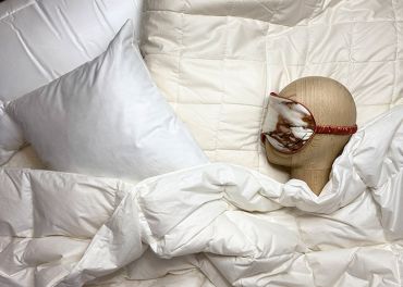 Ein Puppenkopf mit Schlafmaske ruht auf einem Bett, das mit einer weißen Bettdecke und einem Kissen bedeckt ist. Die Umgebung vermittelt eine gemütliche und erholsame Atmosphäre.