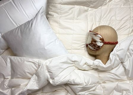 Ein Puppenkopf mit Schlafmaske ruht auf einem Bett, das mit einer weißen Bettdecke und einem Kissen bedeckt ist. Die Umgebung vermittelt eine gemütliche und erholsame Atmosphäre.