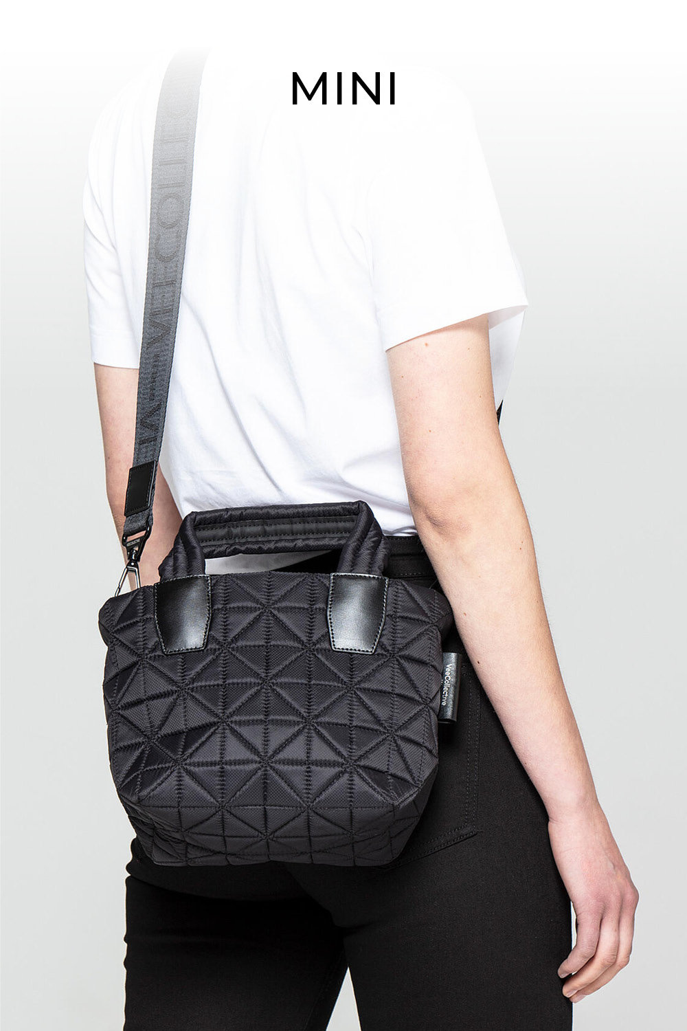 Eine Person in weißem T-Shirt und schwarzer Hose trägt eine kleine schwarze Stepptasche mit Ledergriffen und Schultergurt. Darüber ist das Wort „MINI“ zu sehen. Der Hintergrund ist hellgrau.