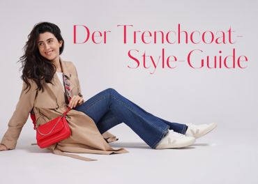 Eine Person mit langen, dunklen Haaren, die einen beigen Trenchcoat, blaue Jeans und weiße Turnschuhe trägt, sitzt auf dem Boden. Sie hat eine rote Handtasche dabei und lächelt. In roter Schrift ist der Text „Der Trenchcoat-Style-Guide“ zu sehen.