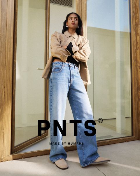 Eine Frau steht selbstbewusst vor einer Glastür und trägt eine beigefarbene Jacke, ein schwarzes Oberteil, weite Bluejeans und beigefarbene Schuhe. Der Text "PNTS MADE BY HUMANS" wird am unteren Rand des Bildes angezeigt.