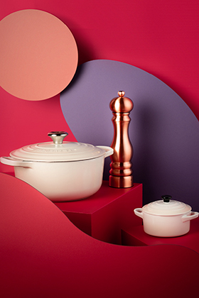 Keramik-Kochgeschirr-Set mit geometrischen Formen in Rot und Lila. Enthält einen großen weißen Topf mit Deckel, einen kleinen weißen Topf mit schwarzem Knauf und eine kupferfarbene Pfeffermühle. Der Hintergrund zeigt kreisförmige Elemente in kräftigen Rot- und Pfirsichfarben.