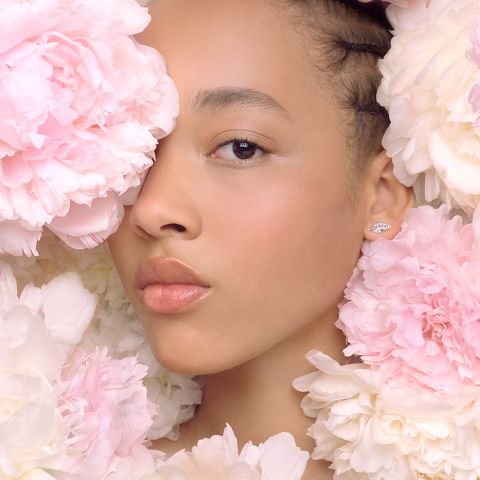 Eine Frau ist teilweise von großen rosa und weißen Pfingstrosen umgeben. Ihr Gesicht ist sanft beleuchtet und zeigt natürliches Make-up und glänzende Lippen. An ihrem sichtbaren Ohr trägt sie einen kleinen, eleganten Ohrring. Der Gesamtton des Bildes ist sanft und heiter.