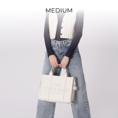Person hält eine weiße Einkaufstasche mit dem Text „THE TOTE BAG“; trägt ein schwarzes Langarmshirt, eine weiße Weste und locker sitzende Jeans. Darüber steht das Wort „MEDIUM“, das die Größe der Tasche angibt.