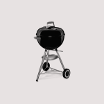 Ein schwarzer, runder Holzkohlegrill im Kesselstil auf Rädern. Der Grill ist auf einem Dreibeinständer mit einer unteren Metallablage und zwei großen Rädern für die Mobilität montiert. Der Deckel hat einen Griff und eine Entlüftung. Der Hintergrund ist schlicht hellgrau.
