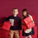 Ein lächelnder Junge und ein lächelndes Mädchen stehen vor einem roten Hintergrund und halten große rote Geschenkboxen mit großen Schleifen in den Händen. Beide tragen festliche Kleidung und hinter ihnen sind weitere verpackte Geschenke gestapelt.