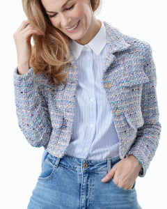 Lächelnde Frau mit langen blonden Haaren, die ein hellblaues gestreiftes Hemd, eine strukturierte helle Jacke und blaue Cargo-Jeans trägt, posiert vor einem hellblauen Hintergrund. Sie berührt sanft ihr Haar mit einer Hand und hat die andere Hand in der Tasche.