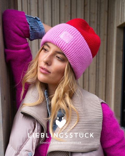 Eine Frau mit einer rosa-roten Strickmütze, einem hellrosa Pullover, einer beigen Pufferweste und einem Jeanshemd steht vor einer Holzwand. Sie sieht nachdenklich aus und hat den Kopf auf ihre Hand gestützt. Der Text lautet "LIEBLINGSSTÜCK.