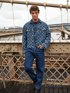 Ein Mann in einer gemusterten Jeansjacke und blauen Jeans steht auf einer Brücke mit Metallgeländer. Im Hintergrund sind ein Fluss und eine weitere Brücke an einem bewölkten Tag zu sehen.