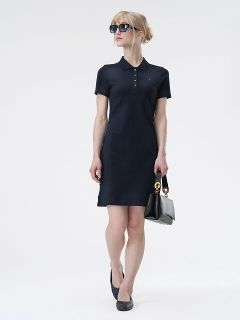 Eine Person mit Sonnenbrille und marineblauem Polokleid steht vor einem schlichten weißen Hintergrund. Sie hält eine schwarze Handtasche und trägt schwarze Ballerinas. Ihre Haare sind hochgesteckt.