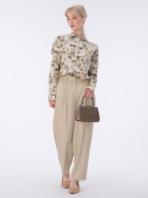 Eine Person in einer geblümten Bluse und beiger Hose steht da und hält eine kleine beige Handtasche in der Hand. Sie hat kurze blonde Haare und trägt flache beige Schuhe. Der Hintergrund ist schlicht weiß.