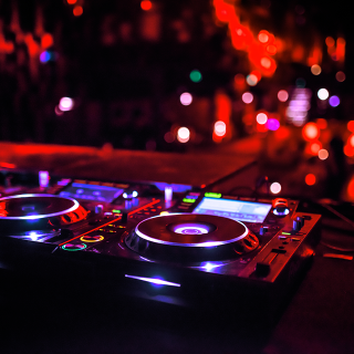 Nahaufnahme eines DJ-Mischpults mit zwei Plattenspielern in einer schwach beleuchteten Nachtclubumgebung. Farbige Lichter und Bokeh-Effekte erzeugen eine lebendige Atmosphäre und werfen rote und violette Farbtöne auf die Geräte.