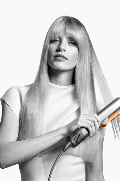 Frau mit langen blonden Haaren, die ein Glätteisen benutzt, ein weißes Oberteil trägt und nach vorne blickt. Kontrastreiches Schwarzweißbild.