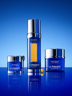 Drei blaue Hautpflegeprodukte von La Prairie, darunter Cremes und ein Serum mit Goldpartikeln, präsentiert auf einer spiegelnden blauen Oberfläche vor blauem Hintergrund.
