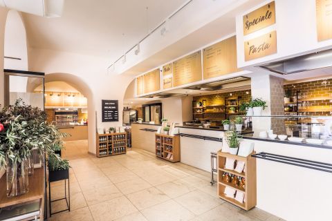 Ein helles, modernes Café-Interieur mit weißen Wänden und gefliestem Boden. Es verfügt über helle Holzregale mit Glasgefäßen, Grünpflanzen und einer Theke mit Menütafeln darüber. Warmes Licht erhellt den Raum und schafft eine einladende Atmosphäre.
