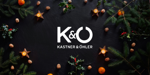Festliches Weihnachtsflachbild mit Stechpalmen, immergrünen Zweigen, Walnüssen, sternförmigen Kerzen, Orangen und roten Beeren. In der Mitte ist das Logo „Kastner & Öhler“ auf dunklem Hintergrund zu sehen.
