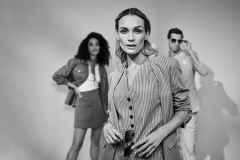 Im Vordergrund steht eine selbstbewusste Frau im gestreiften Outfit mit Sonnenbrille. Dahinter posieren eine Frau und ein Mann stilvoll, wobei der Mann seine Sonnenbrille zurechtrückt. Das Bild ist schwarz-weiß.