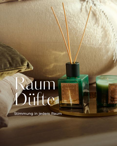 Ein grüner Rohrdiffusor und eine Kerze mit goldenen Etiketten stehen auf einem goldenen Tablett, daneben ein beiges Kissen und ein olivgrünes Kissen. Der Text lautet: "Raum Düfte. Stimmung in jedem Raum.