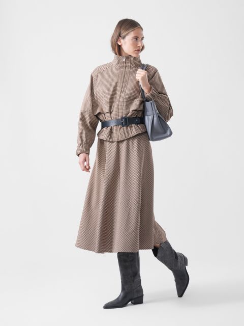 Eine Frau in einem braunen, gemusterten Rock und einer Jacke mit schwarzem Gürtel, grauen Stiefeln und einer grauen Handtasche, die vor einem schlichten weißen Hintergrund steht.