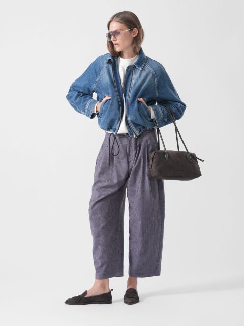 Eine Person, die eine Jeansjacke, eine weite graue Hose, Slipper und eine Sonnenbrille trägt sowie eine schwarze Handtasche bei sich hat, steht vor einem schlichten weißen Hintergrund.