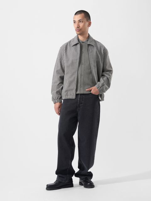 Vor einem schlichten weißen Hintergrund steht ein Mann, der eine graue Karojacke, ein graues Hemd, weite schwarze Jeans und schwarze Schuhe trägt.