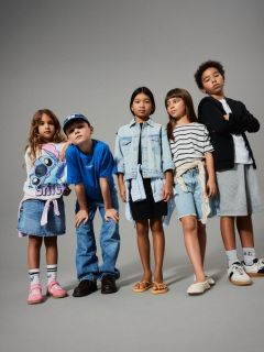 Ein kleines Kind mit blondem Haar steht lächelnd vor einem grünen Hintergrund und trägt einen braunen ärmellosen Pullover über einem geblümten Kleid, kniehohe Socken und braune Schuhe.