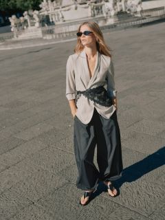 Eine Frau in einem braunen, übergroßen Anzug mit schwarzem Gürtel und Goldschmuck geht selbstbewusst über eine Straße in der Stadt und trägt eine große schwarze Handtasche. Im Hintergrund sind sitzende Café-Besucher zu sehen.