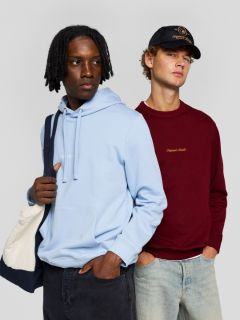 Ein junger Mann mit kurzen braunen Haaren, der ein cremefarbenes T-Shirt mit marineblauem Aufdruck auf dem Rücken, hellblaue Jeans und beigefarbene Turnschuhe trägt, sitzt auf dem Boden und schaut über die Schulter in die Kamera.