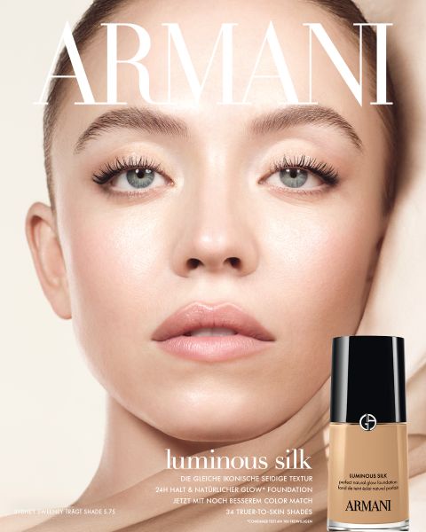 Nahaufnahme eines Modells mit heller Haut und blauen Augen, das eine taufrische, strahlende Haut zeigt. In der unteren rechten Ecke ist eine Tube Armani-Hautfarbe abgebildet, deren Text für 24-Stunden-Hydratation und 20 strahlende Farbtöne wirbt.