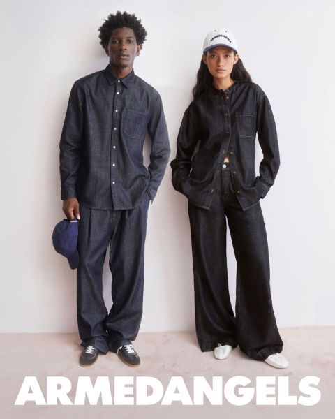 Zwei junge Erwachsene in Jeanskleidung stehen zusammen im Freien, einer mit erhobenem Arm, der andere mit einem Koffer, der mit Aufklebern bedeckt ist. Am unteren Rand steht in fetter weißer Schrift "ARMEDANGELS".