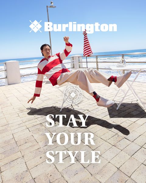 Ein Mann und eine Frau in stilvollen Outfits posieren selbstbewusst auf einer weißen Bank vor einer grünen Wand mit den Texten "Burlington" und "Stay Your Style" über und unter ihnen.